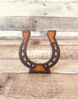 The Faire Horseshoe Rustic Western Doodad is a brown metal tabletop figurine with a vintage look, standing upright on wood for charming rustic decor.