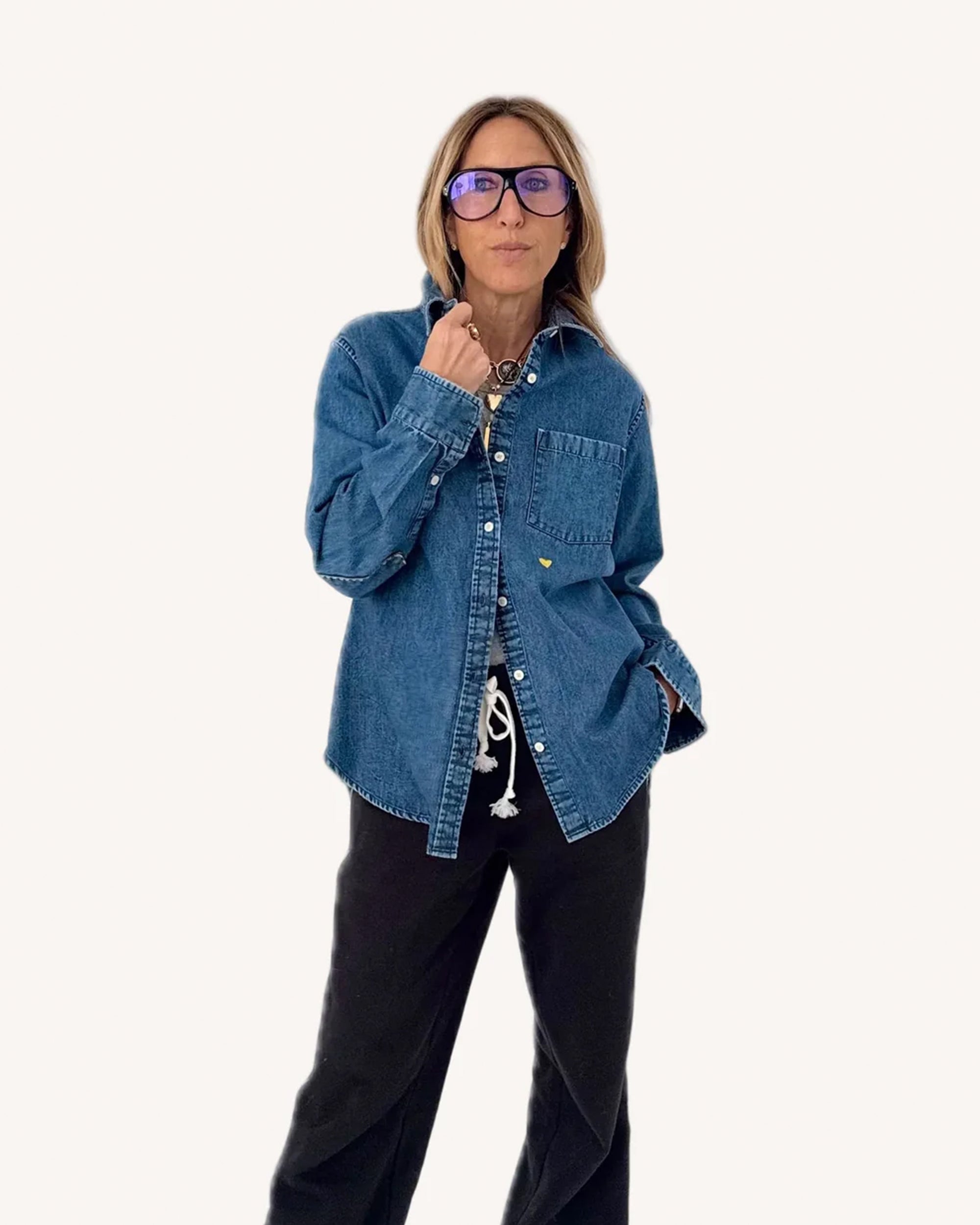 A person models the oversized-fit Mia Denim Shirt by Kerri Rosenthal, featuring an embroidered heart, paired with black drawstring pants and large dark-rimmed glasses, posing confidently against a plain white background.