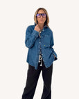 A person models the oversized-fit Mia Denim Shirt by Kerri Rosenthal, featuring an embroidered heart, paired with black drawstring pants and large dark-rimmed glasses, posing confidently against a plain white background.