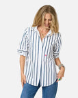 A woman with shoulder-length blonde hair wears the Kerri Rosenthal Mia Mr Sharpie Stripe Shirt, featuring vertical navy stripes, paired with blue jeans and layered jewelry, smiling as she looks down against a white background.
