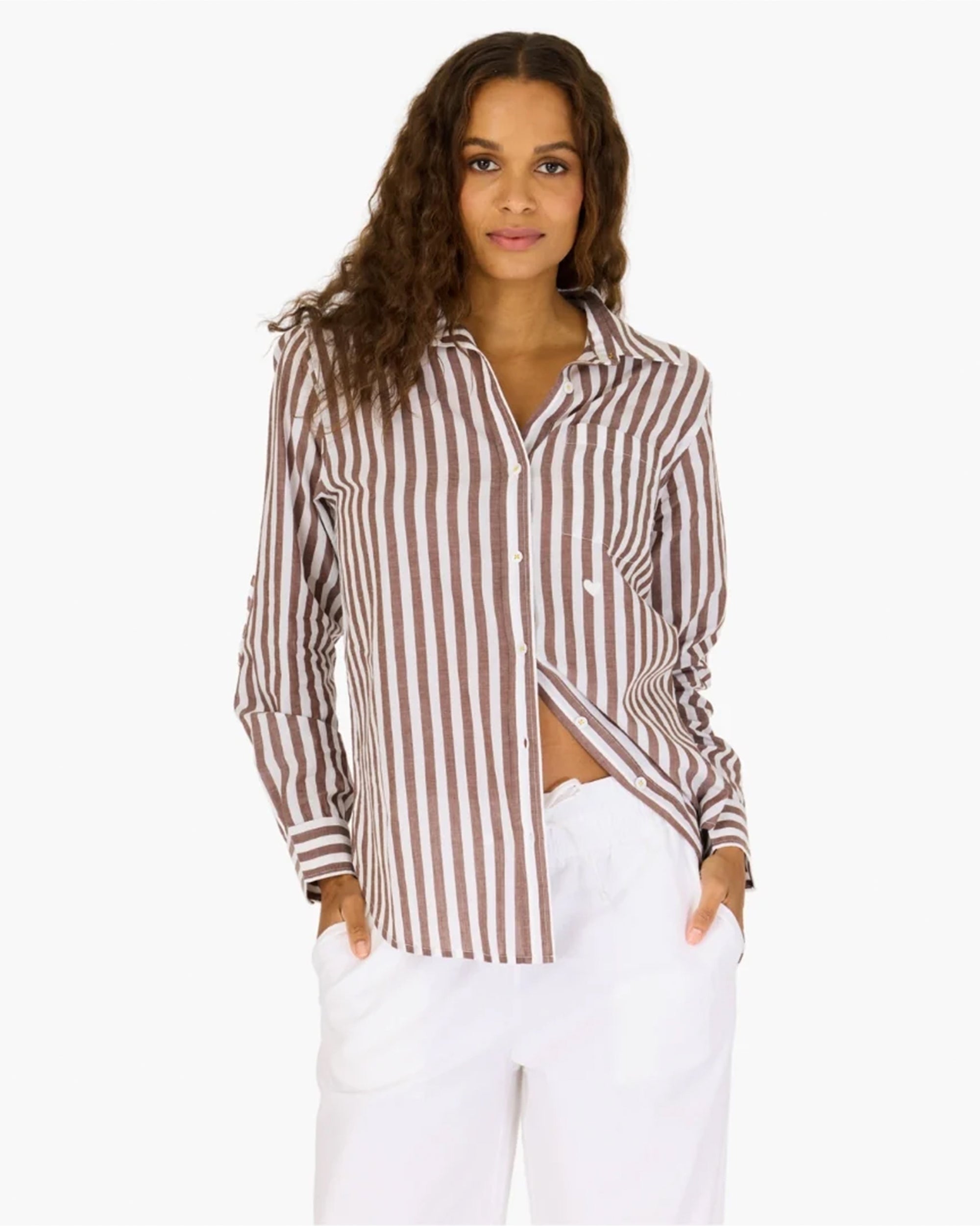 A woman with long, wavy brown hair wears the Mia Stripe Shirt by Kerri Rosenthal and white pants. She stands with her hands in her pockets against a plain white background, smiling softly.