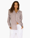 A woman with long, wavy brown hair wears the Mia Stripe Shirt by Kerri Rosenthal and white pants. She stands with her hands in her pockets against a plain white background, smiling softly.