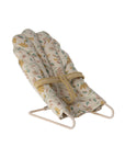 Soft beige Maileg micro babysitter seat with safety strap on a white frame, perfect for dollhouse play.