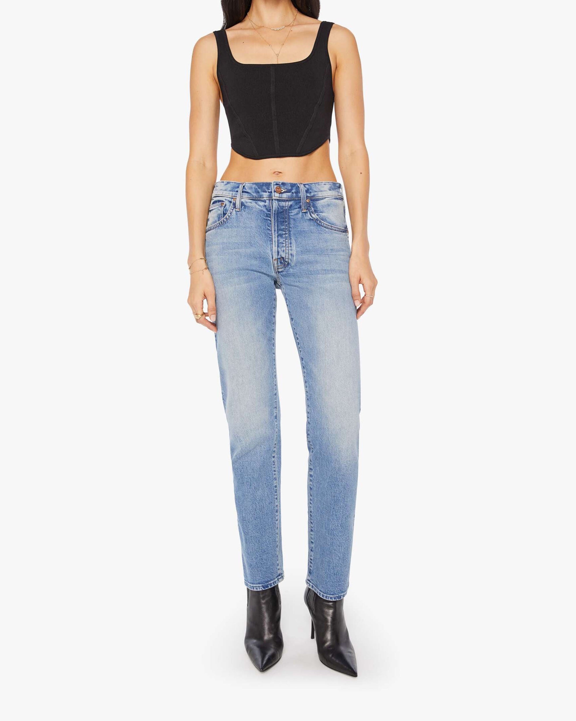 A person in a black cropped tank top, Mother Mid Rise Hiker Hover light blue stretch denim slim straight-leg jeans, and pointed black ankle boots stands against a plain white background.
