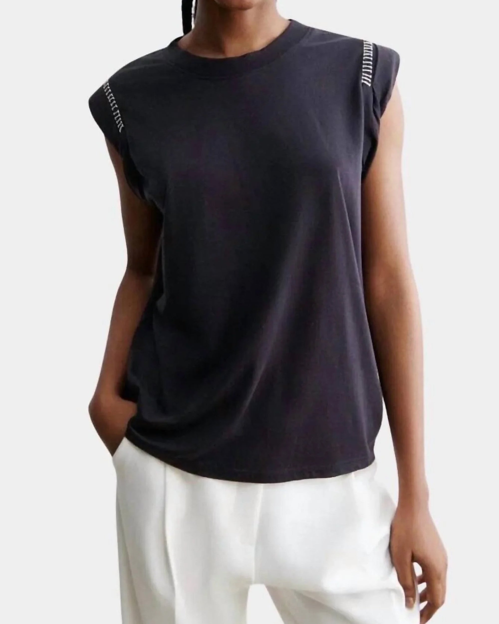 A person wearing the Rachel Comey Miles Tee with white text on the shoulders and loose-fitting white pants stands with one hand in a pocket against a plain background.