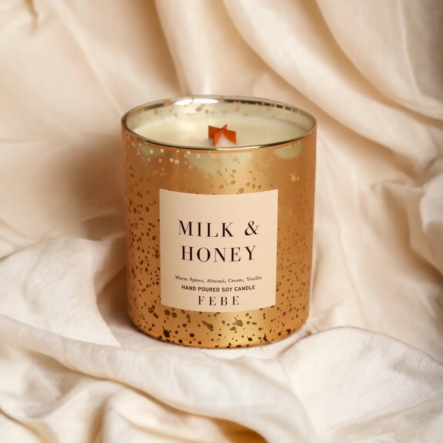 A FEBE candle labeled &quot;Milk &amp; Honey&quot; in a speckled gold container, resting on a soft, wrinkled cream fabric background in Arizona style.