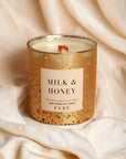 A FEBE candle labeled "Milk & Honey" in a speckled gold container, resting on a soft, wrinkled cream fabric background in Arizona style.