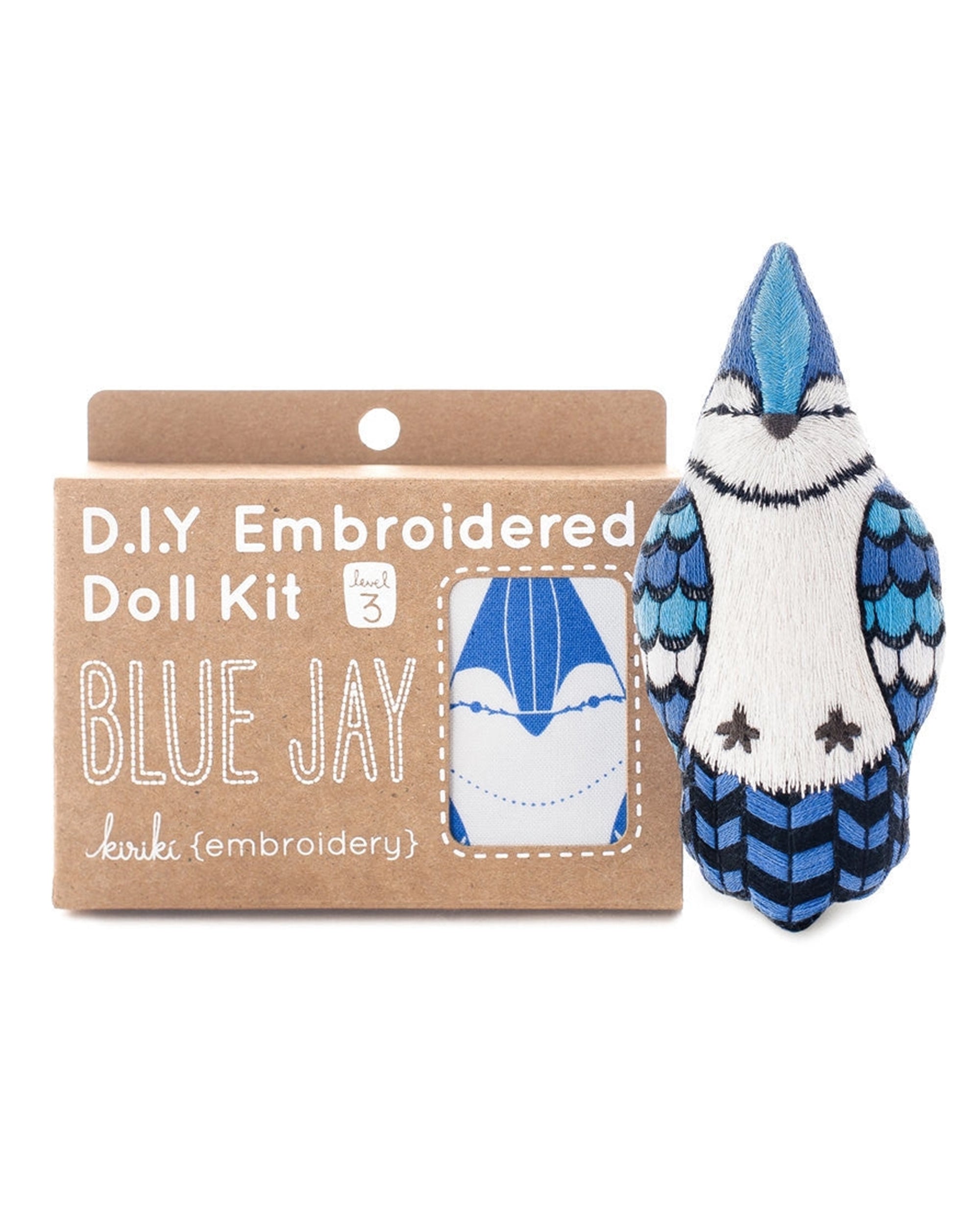 The Faire Blue Jay Embroidery Kit comes in a brown package with a blue jay design. Next to it, a finished plush blue jay doll is sewn in blue, white, and black.