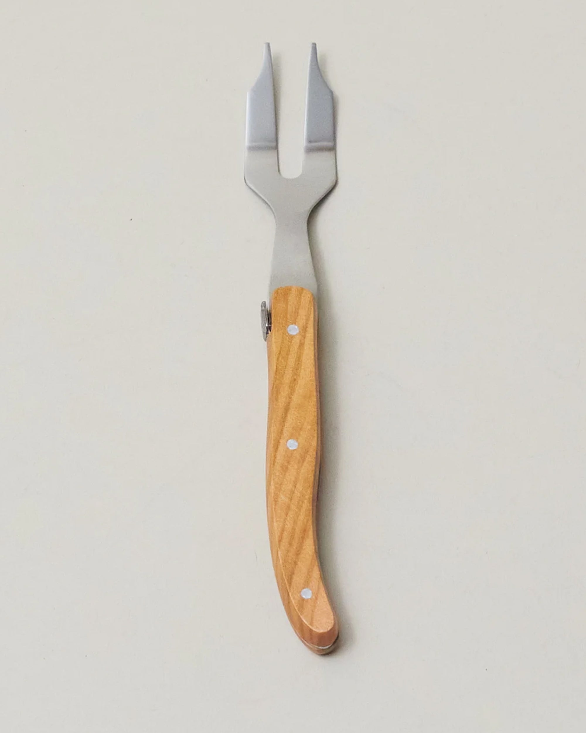 The Be Home Laguiole Mini Fork, featuring a wooden handle and stainless steel prongs inspired by French craftsmanship, rests on a plain, light-colored surface.