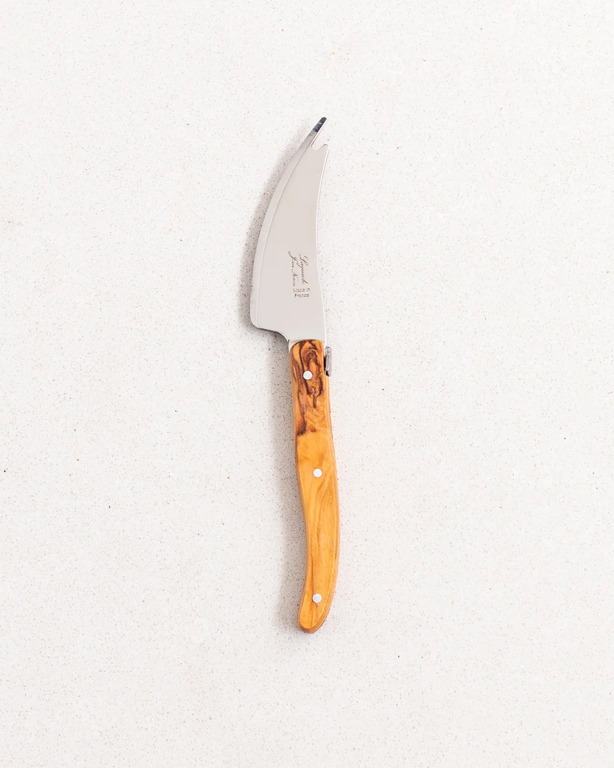 The Be Home Laguiole Mini Pronged Knife, featuring a curved stainless steel blade and light brown wooden handle, exemplifies French craftsmanship and rests on a light speckled surface.