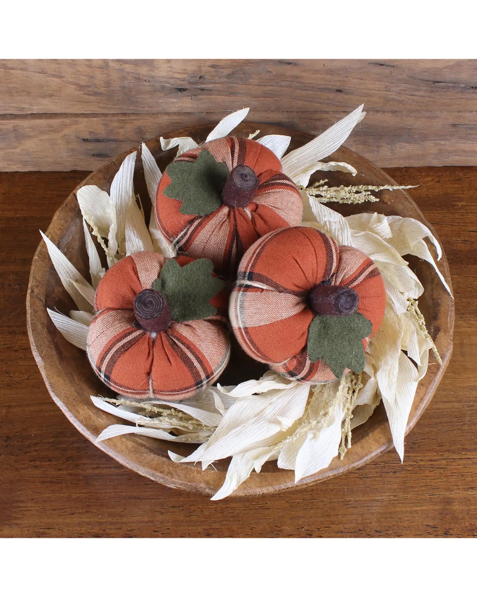 A wooden bowl displays three Faire Mini Pumpkin Plaid decorations with orange plaid designs, felt leaves, and brown stems, surrounded by dried corn husks on wood—a charming choice for cozy autumn decor.