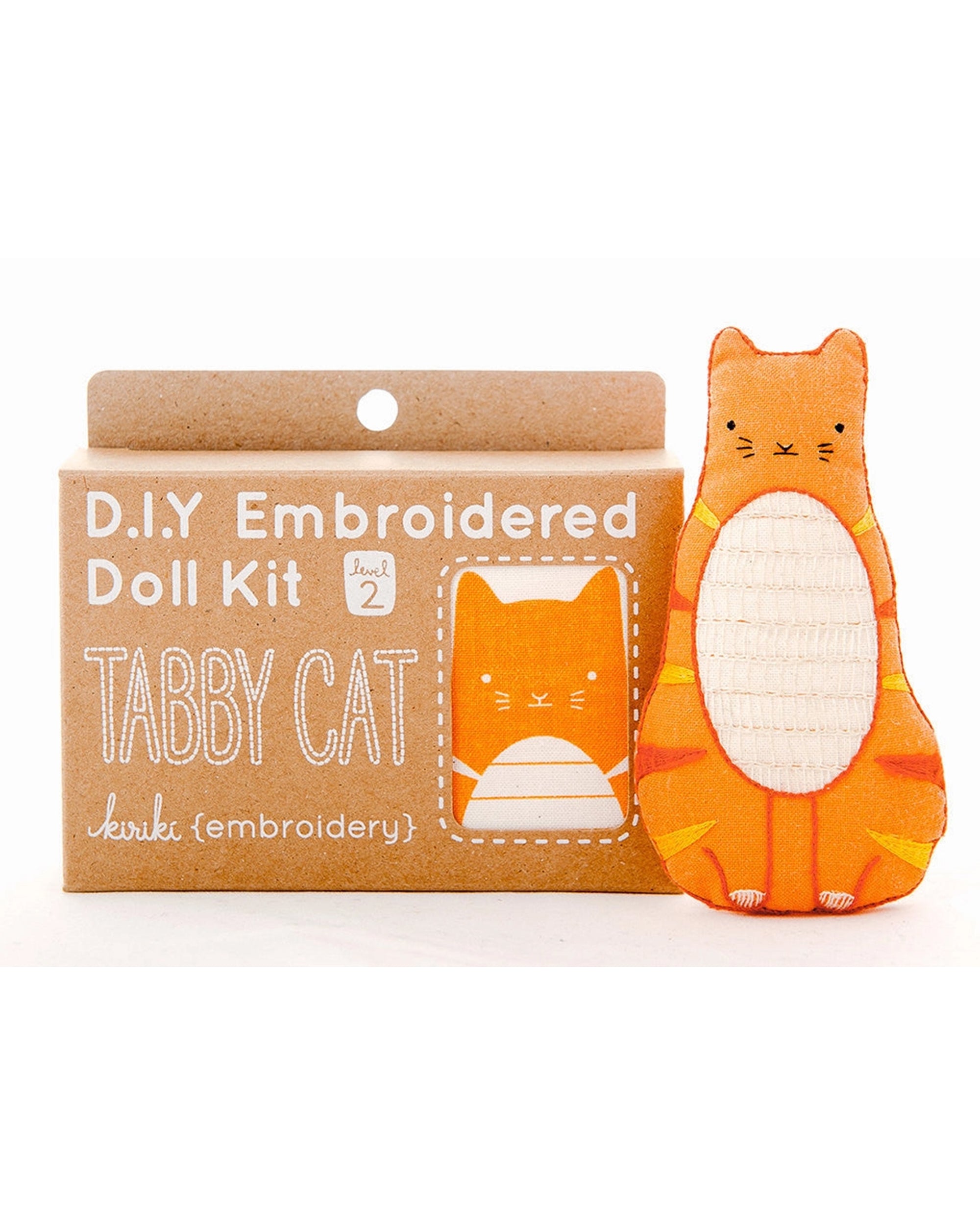The Faire Tabby Cat Embroidery Kit is shown in its brown box with white text and a cat illustration. Beside it sits a completed orange tabby doll, hand-stitched from screen-printed fabric with charming details.