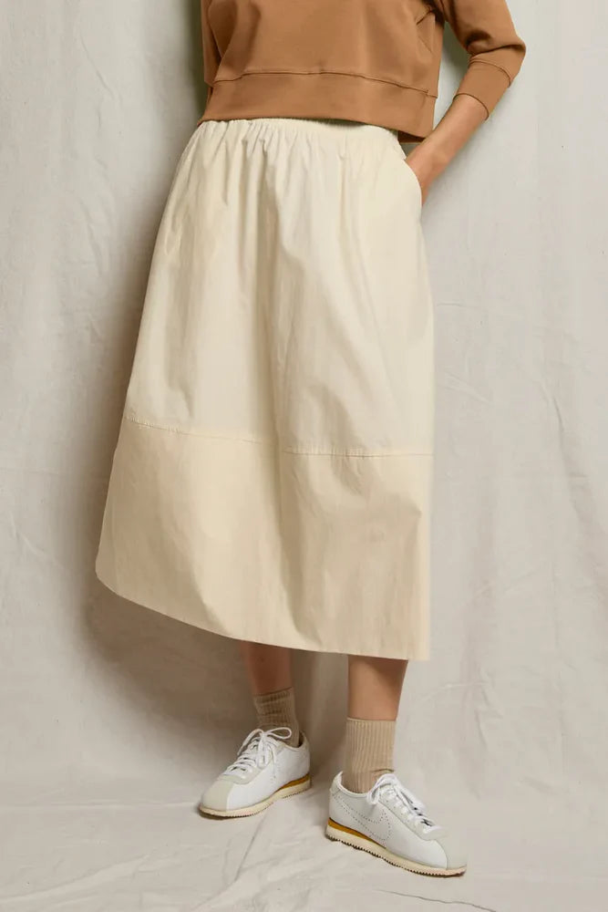 A person wears the Perfectwhitetee Poplin A Line Skirt with pockets, paired with a tan sweatshirt, beige socks, and white sneakers. They stand against a light fabric backdrop with hands casually tucked in the skirt’s pockets.