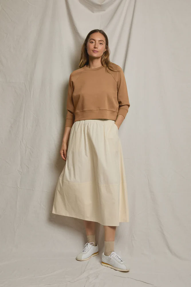 A woman with long hair and a neutral expression stands before a beige backdrop, wearing a light brown sweatshirt, beige socks, white sneakers, and the Poplin A Line Skirt by Perfectwhitetee in cream.