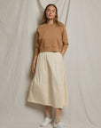 A woman with long hair and a neutral expression stands before a beige backdrop, wearing a light brown sweatshirt, beige socks, white sneakers, and the Poplin A Line Skirt by Perfectwhitetee in cream.