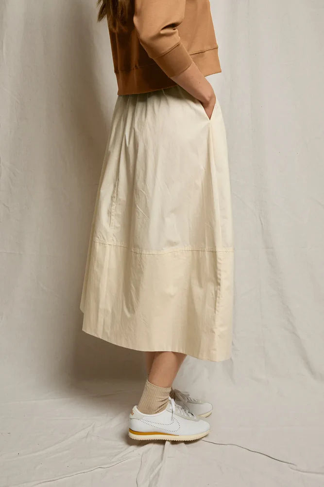 A person stands sideways in a tan sweatshirt, Perfectwhitetee Poplin A Line Skirt in cream with pockets, beige ankle socks, and white sneakers against a light fabric backdrop.