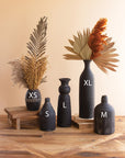 Five Kalalou, Inc Black Clay Vases in sizes XS, S, M, L, and XL with dried plants and leaves are displayed on a wooden surface against a beige background—adding modern style to any decor.