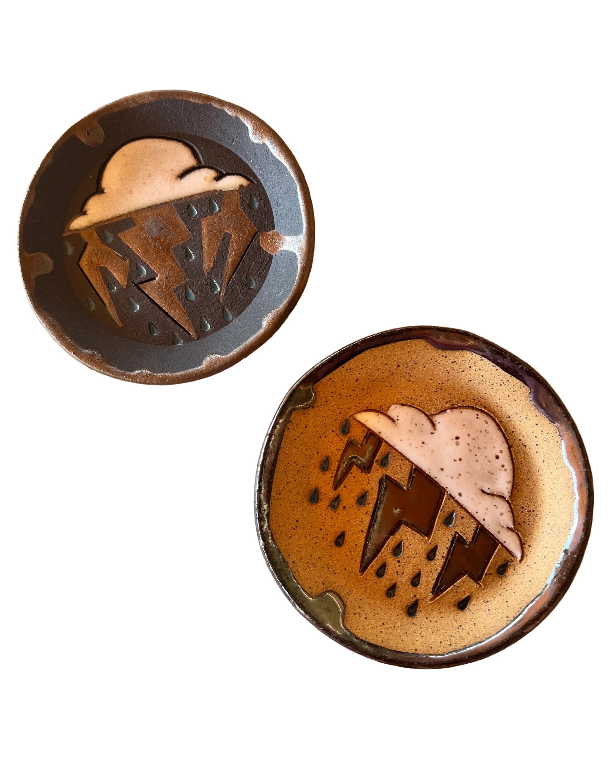 The Faire Monsoon Trinket Dish set includes two round ceramic dishes—one in dark, one in light earthy tones—each with a cloud, lightning bolts, and raindrops. Ideal as monsoon-inspired gifts or handmade desert décor.