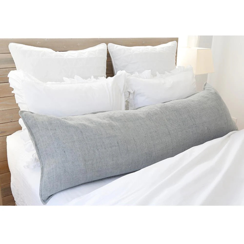 The POM POM Montauk Body Pillow in light gray heavy weight linen rests across a neatly made bed with white sheets and four ruffled white pillows set against a wooden headboard.