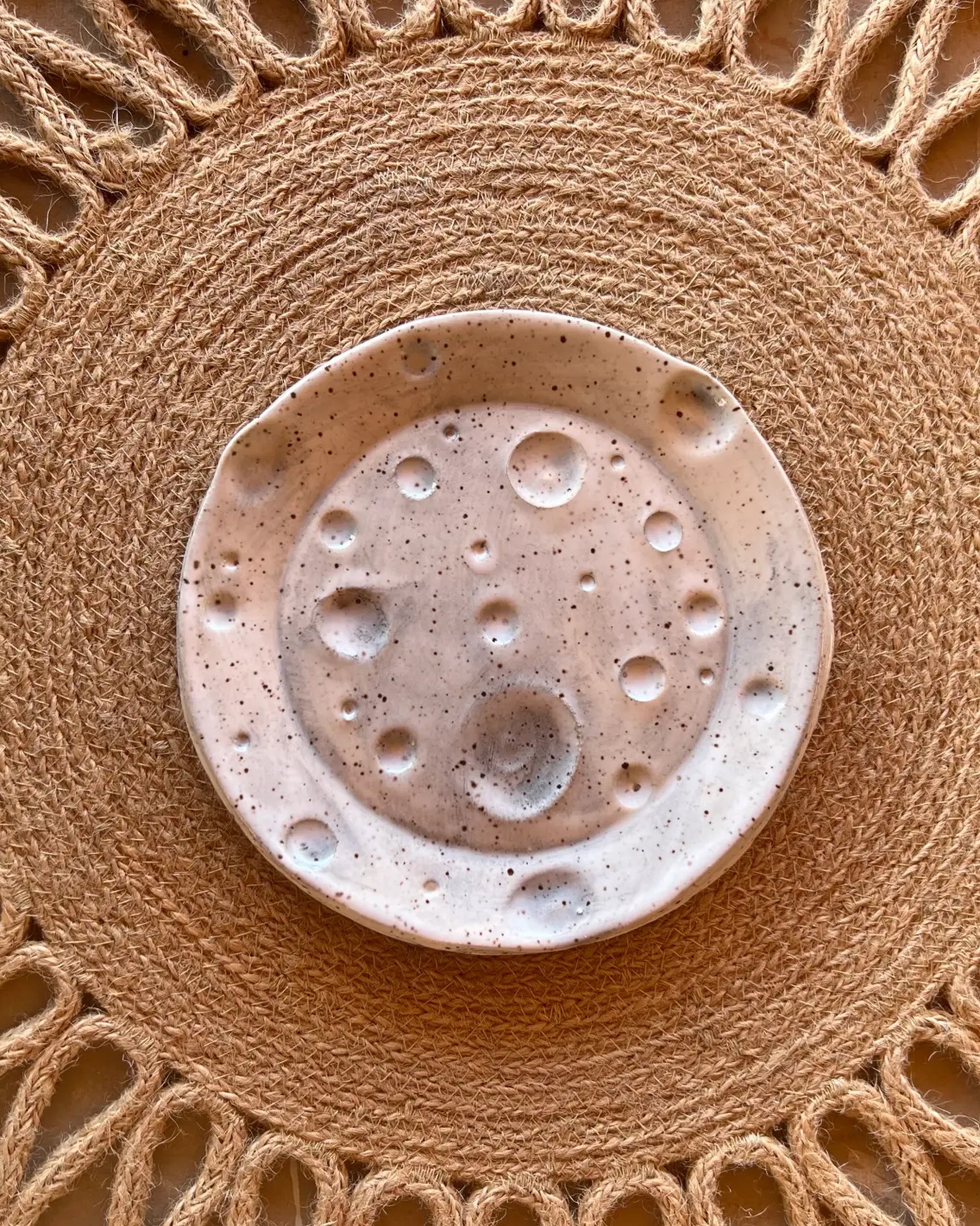 Faire’s Moon Trinkets rest in a ceramic dish featuring a speckled, cratered surface that resembles the moon, displayed on a fringed woven placemat—perfect for showcasing your favorite celestial décor.