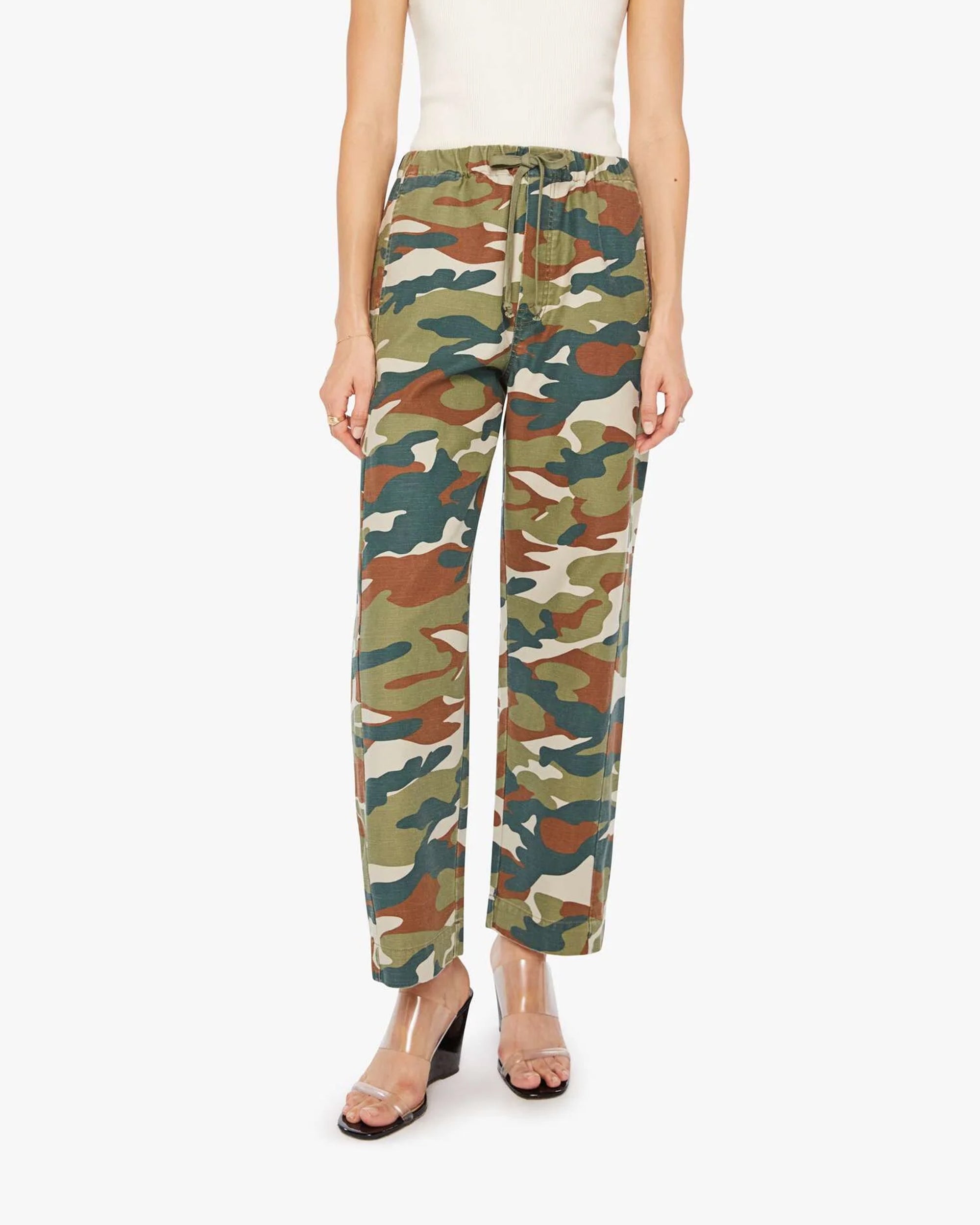 A person wears the Mother "The Drawn Half-Pipe Ankle" high-waisted camouflage pants with a drawstring waist, paired with a white sleeveless top and clear high-heeled sandals.