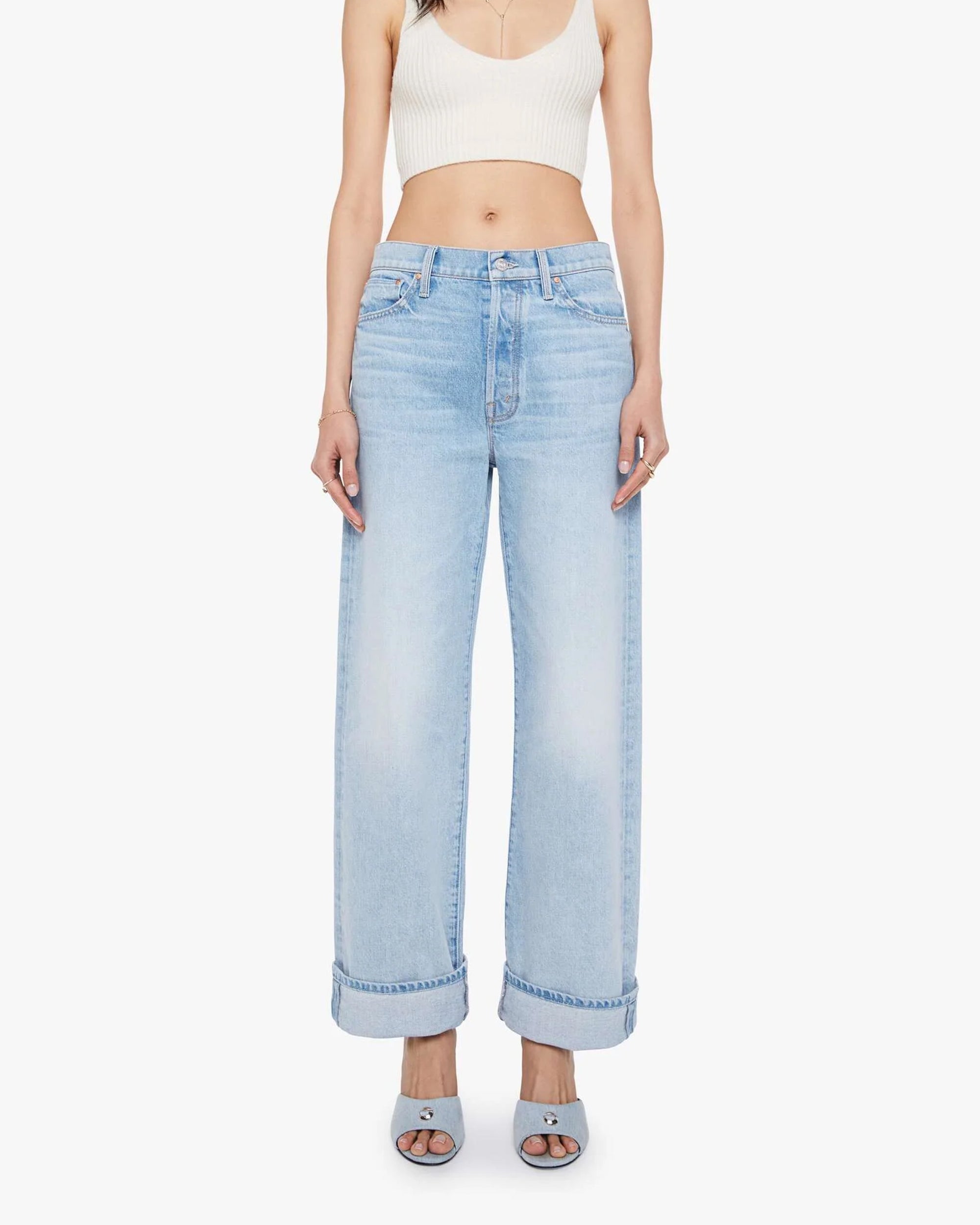 A woman stands against a plain white background wearing The Dodger Button Hover Cuff jeans by Mother, styled with a white cropped tank and light blue open-toe block heel shoes.