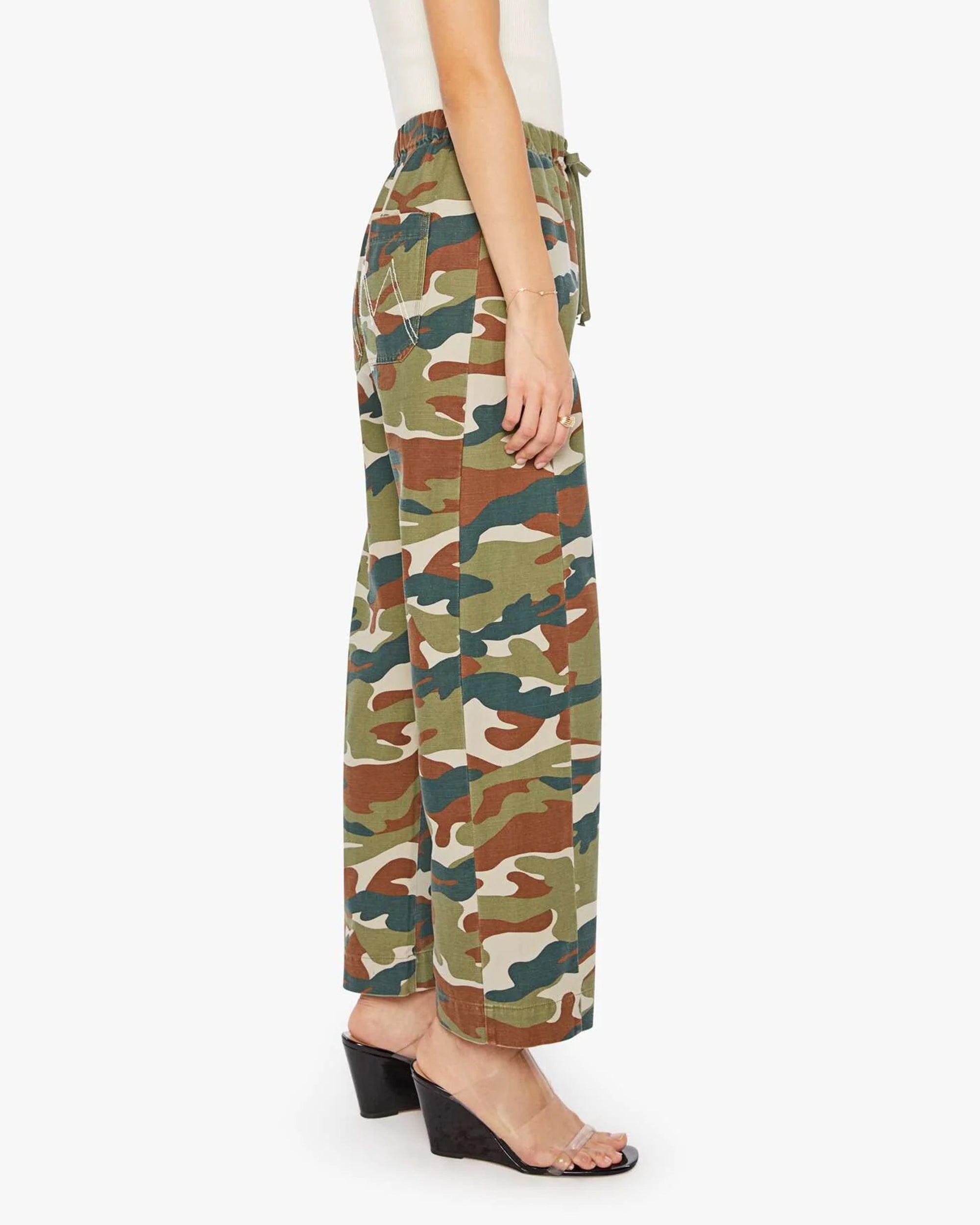 A person wears Mother’s The Drawn Half-Pipe Ankle high-waisted camouflage cotton pants with a drawstring waist, clear strap heels, and a white sleeveless top, shown in profile against a plain background.