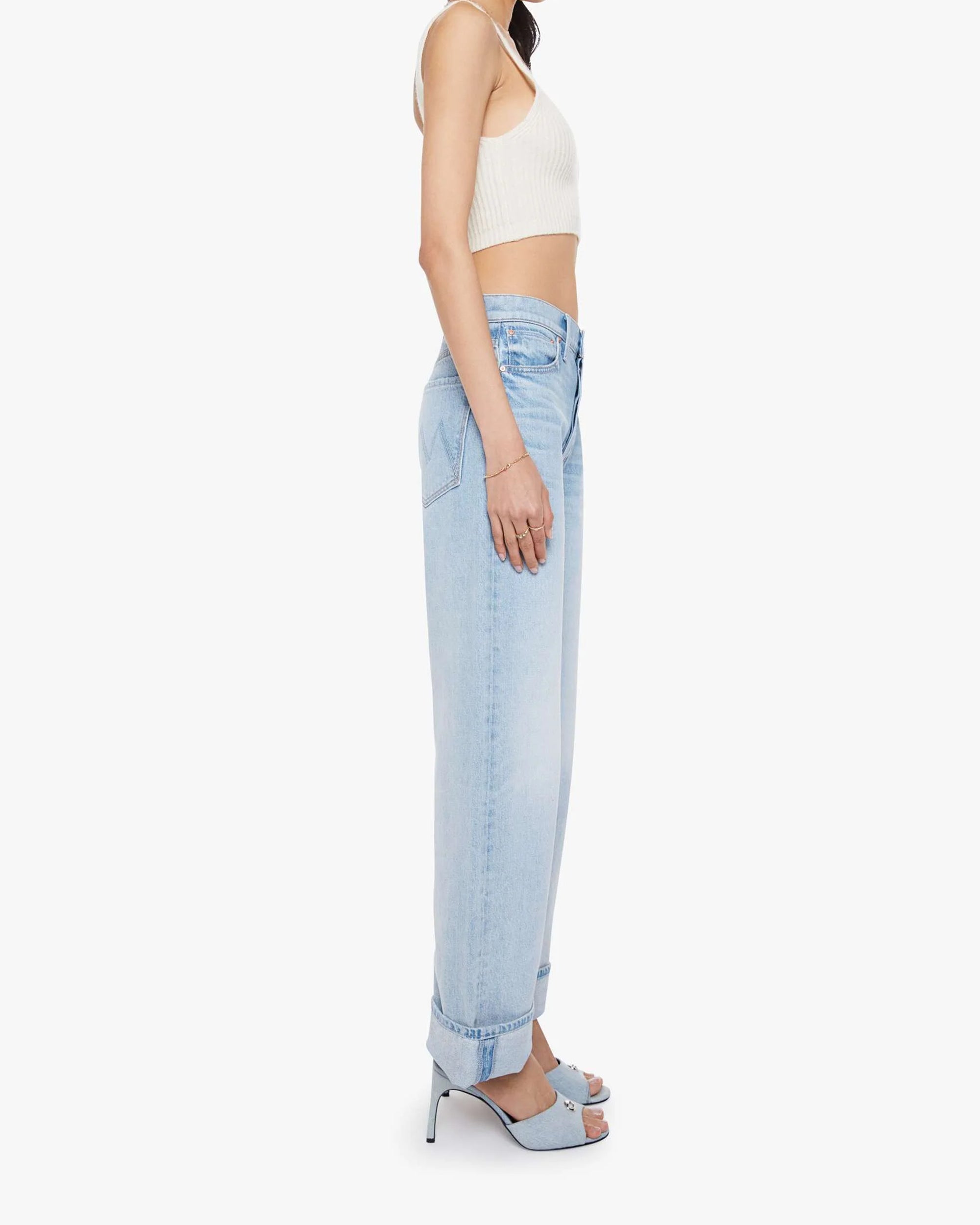A woman in profile wears the Mother The Dodger Button Hover Cuff light blue slouchy jeans with loose straight legs and cuffs, styled with a white sleeveless crop top and blue high-heeled sandals against a plain background.