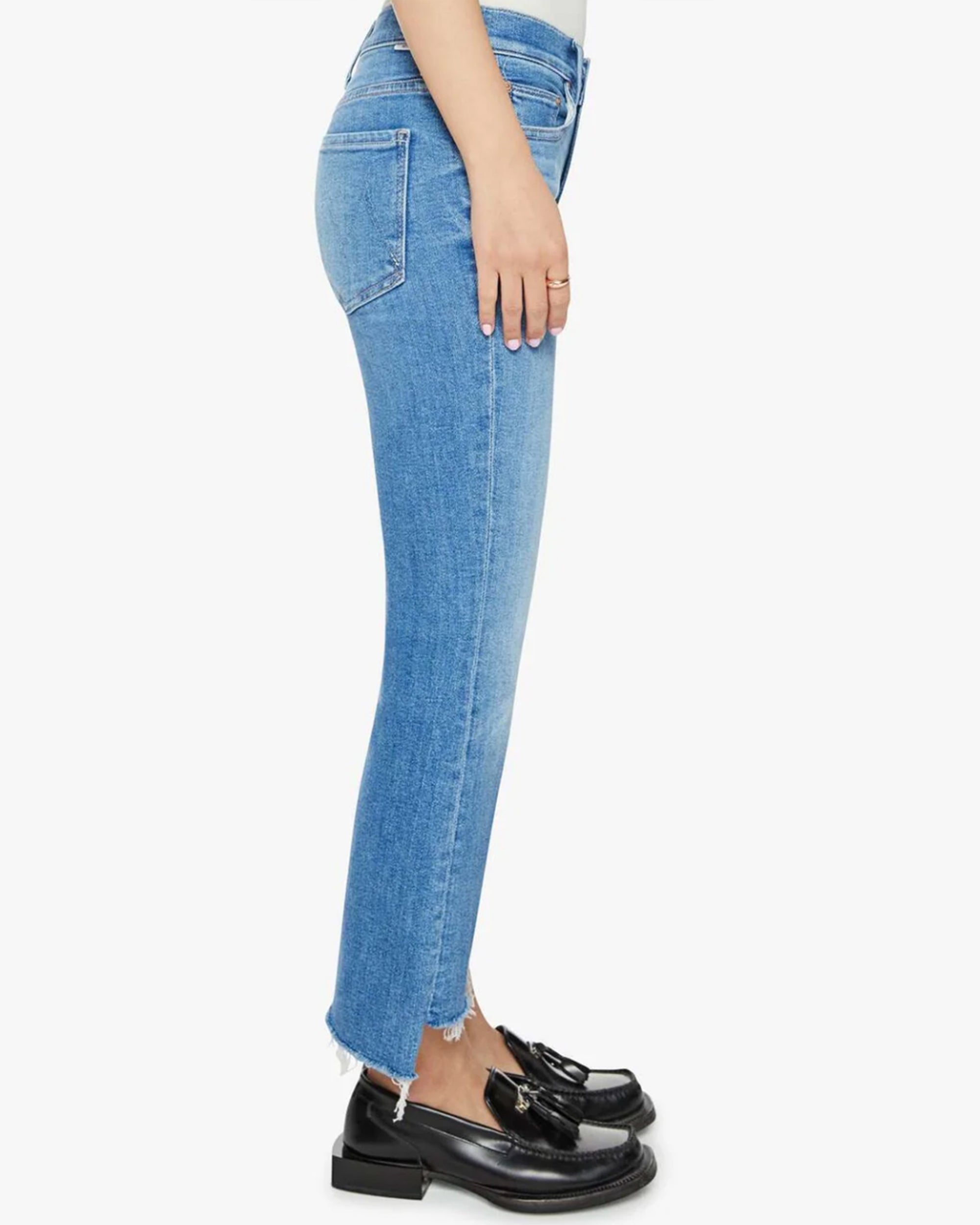 A person standing sideways wears the Mother Lil Insider Crop Step Fray high-waisted bootcut light blue jeans with frayed hems and black tassel loafers, set against a plain white background.