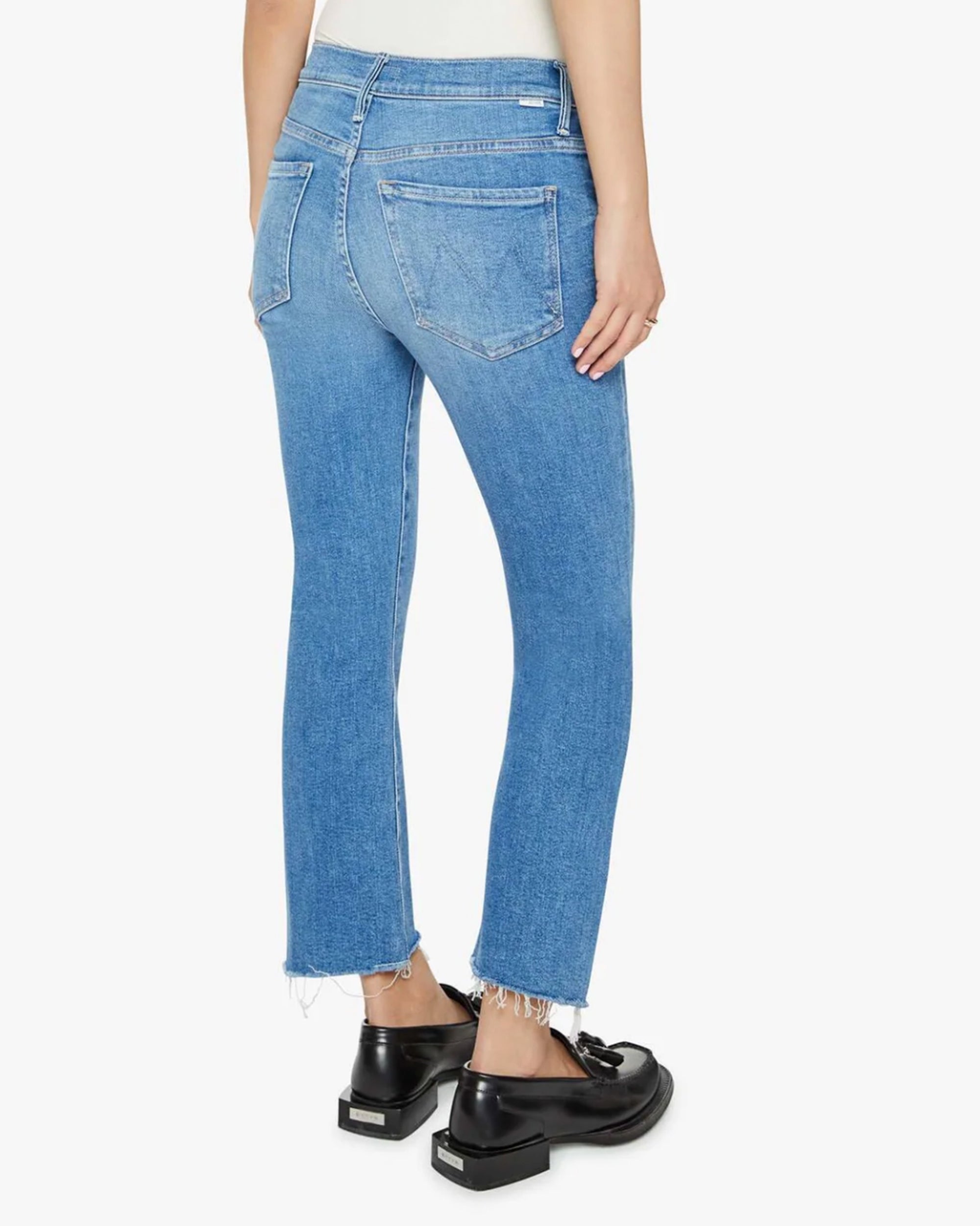 A person is shown from the back, standing against a plain white background, wearing Mother Lil Insider Crop Step Fray high-waisted light blue jeans with a slightly frayed hem and black loafers.