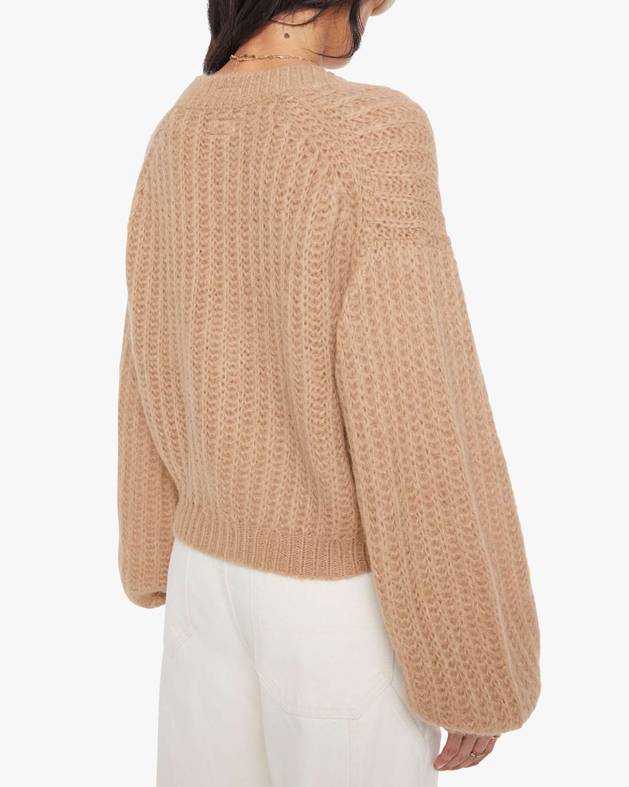 A woman with dark hair models Mother’s The Puffy Sleeve Varsity Cardi—a textured, loose tan sweater with balloon sleeves—paired with high-waisted white pants, shown from the back against a plain white background.