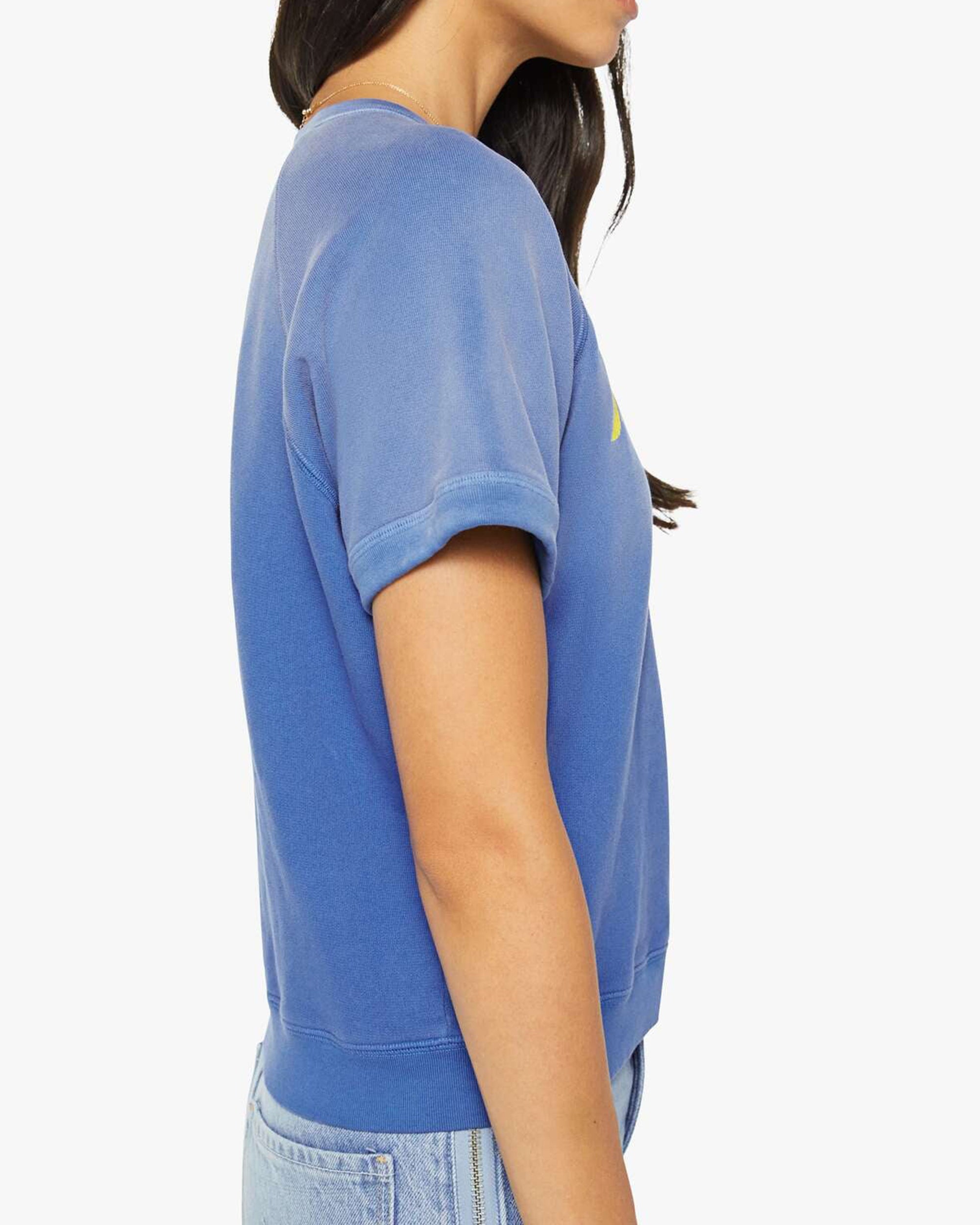 A woman with long dark hair, shown from the side against a white background, wears Mother's The Short Sleeve Concert Tee in blue with rolled sleeves and light blue jeans.