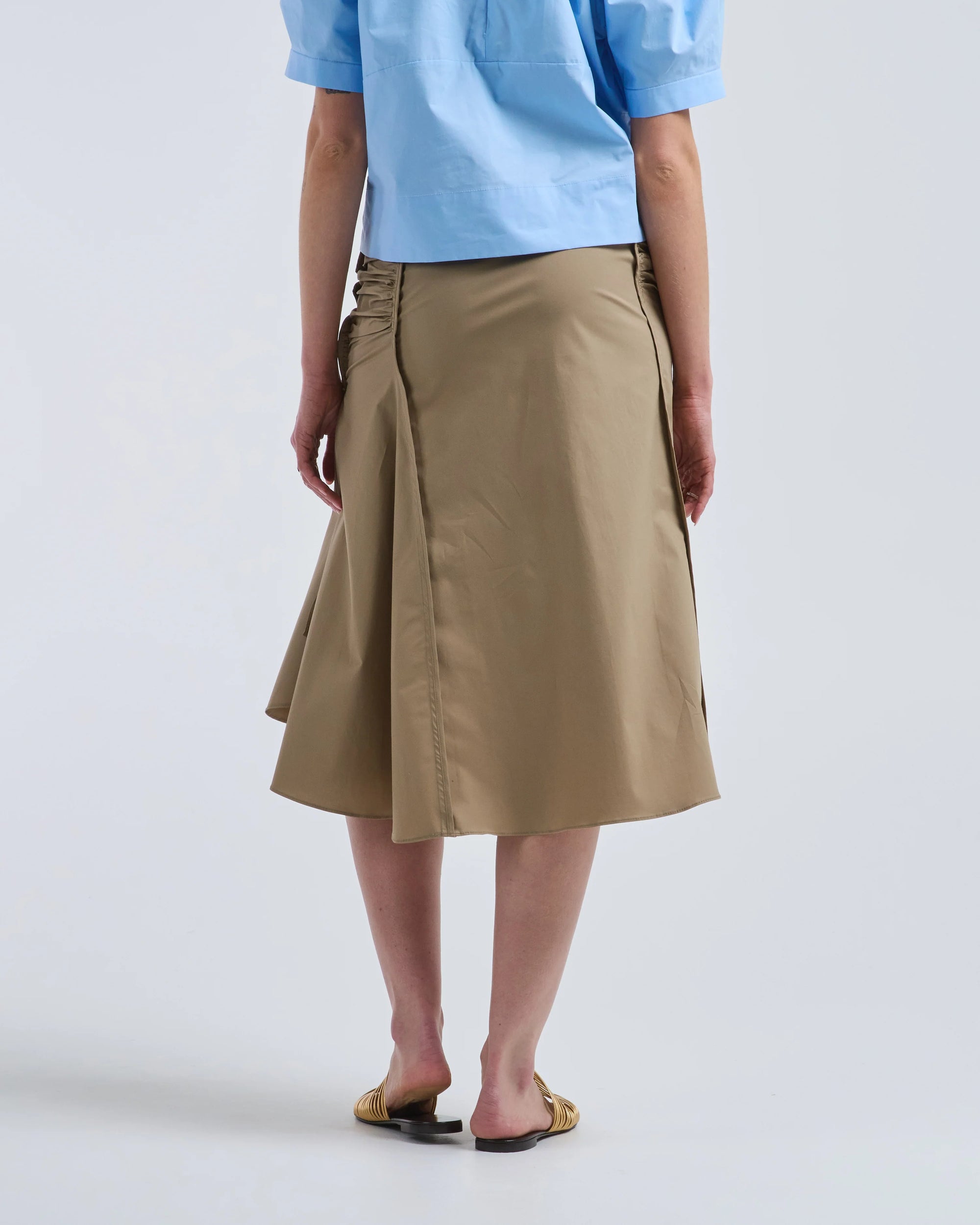 A person stands facing away, wearing a light blue short-sleeve shirt, tan sandals, and the Rachel Comey Muci Skirt—a high waisted khaki skirt with ruched sides—against a plain light background.