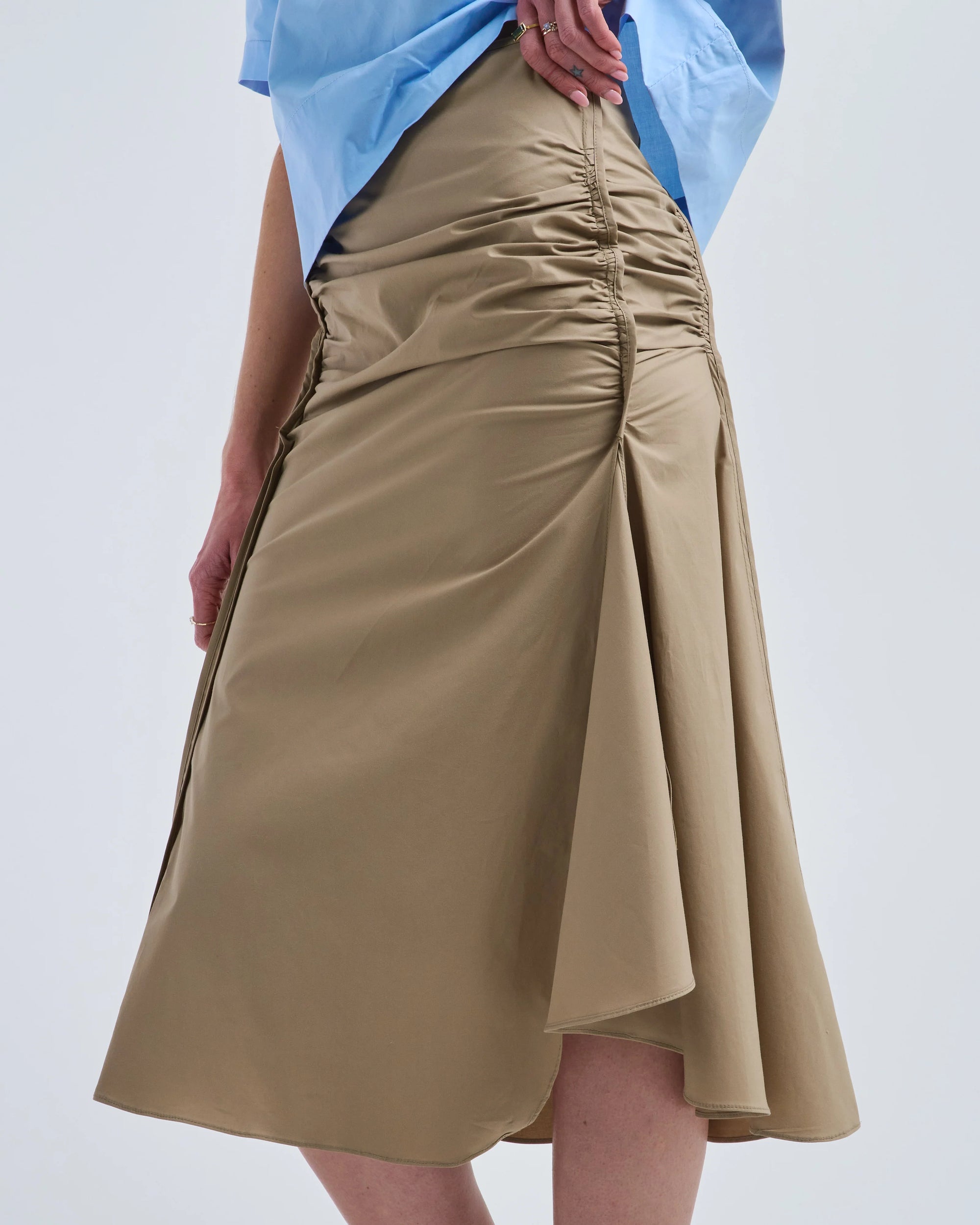 A person wearing a light blue shirt and the Rachel Comey Muci Skirt—a high-waisted, beige ruched knee-length skirt with a front slit—stands against a plain light background.