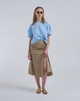 A woman stands against a plain light background wearing sunglasses, a loose light blue T-shirt, gold sandals, and the Rachel Comey Muci Skirt— a high-waisted beige ruched midi skirt with a slit.