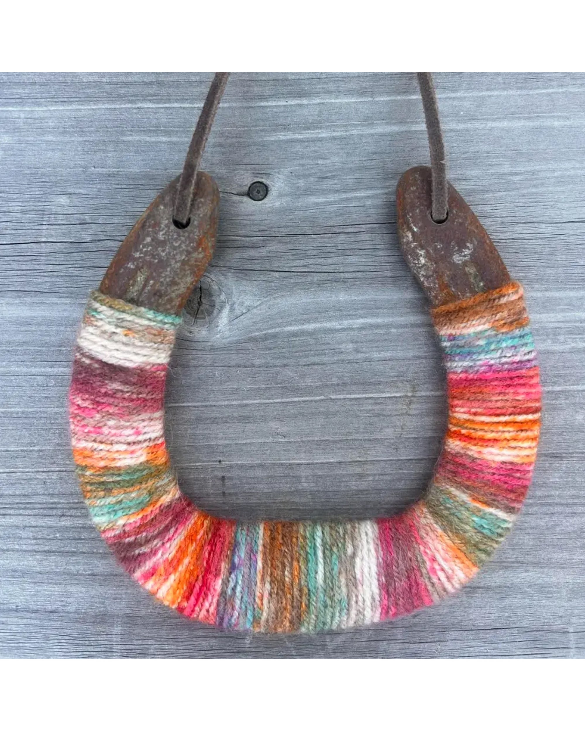 The Faire Lucky Horseshoe features a rustic upcycled horseshoe wrapped in colorful yarn and suspended by a leather cord, making it a unique housewarming gift.
