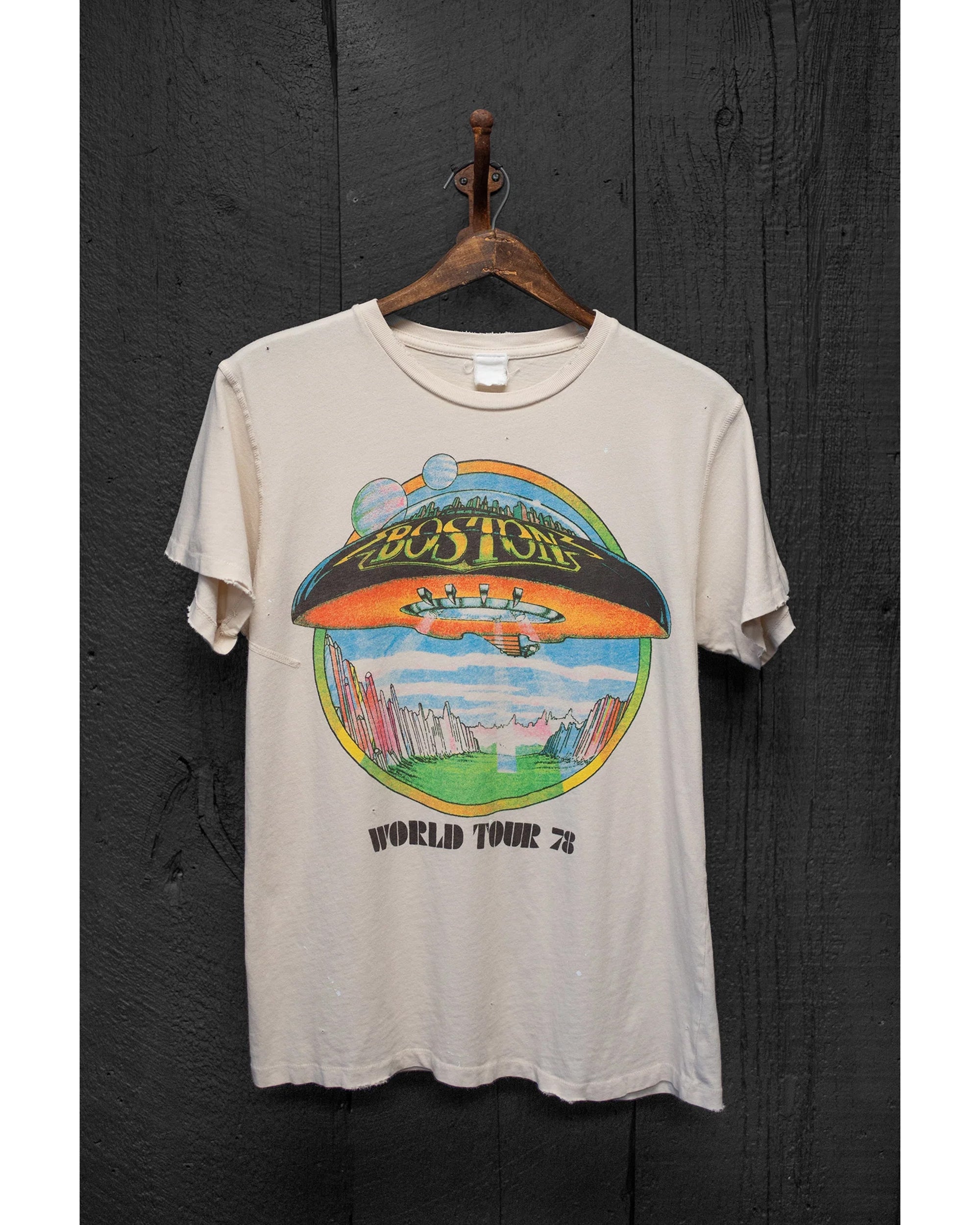The Made Worn Boston Crew Tee, a vintage unisex fit in white, hangs on a wooden hanger against dark wood. It features a colorful "Boston World Tour '78" graphic with a spaceship, planets, and cityscape.