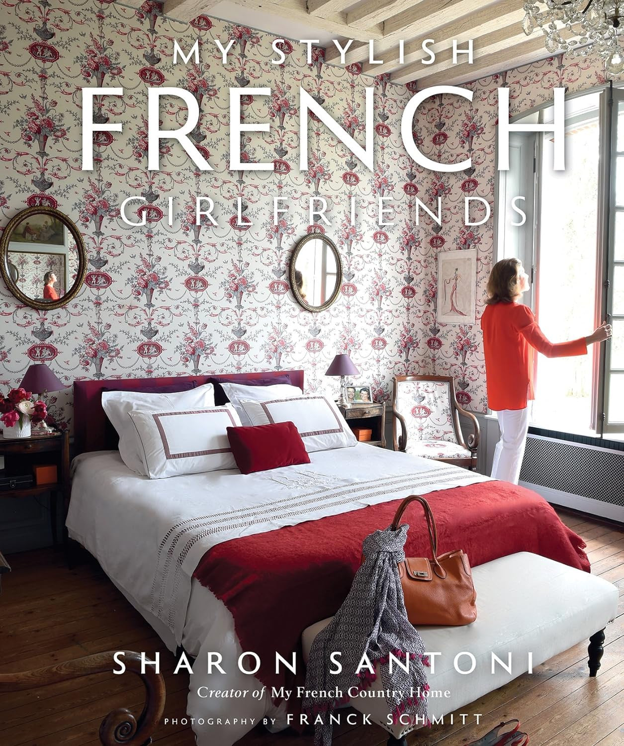 A woman in a stylish bungalow looks out a window beside an elegantly made bed with white linens. The room features ornate wallpaper and wooden flooring. A book cover titled &quot;My Stylish French Girlfriends&quot; published by Gibbs Smith Publisher is displayed.