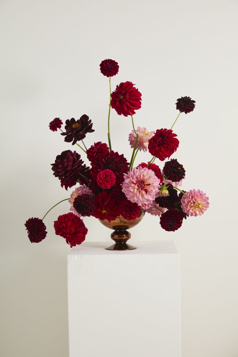 The Naomi Compote by Accent Decor displays dark red, bright red, and light pink dahlias with long stems in a brown vase, set on a white pedestal against a plain white background.