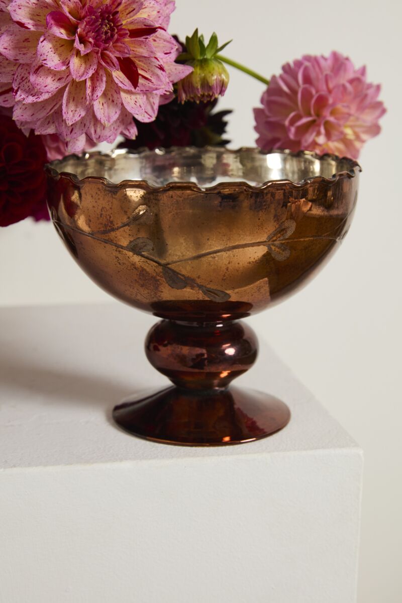 The Naomi Compote by Accent Decor features bronze-colored mercury glass with a scalloped rim, set on a white pedestal and filled with pink and purple dahlias.