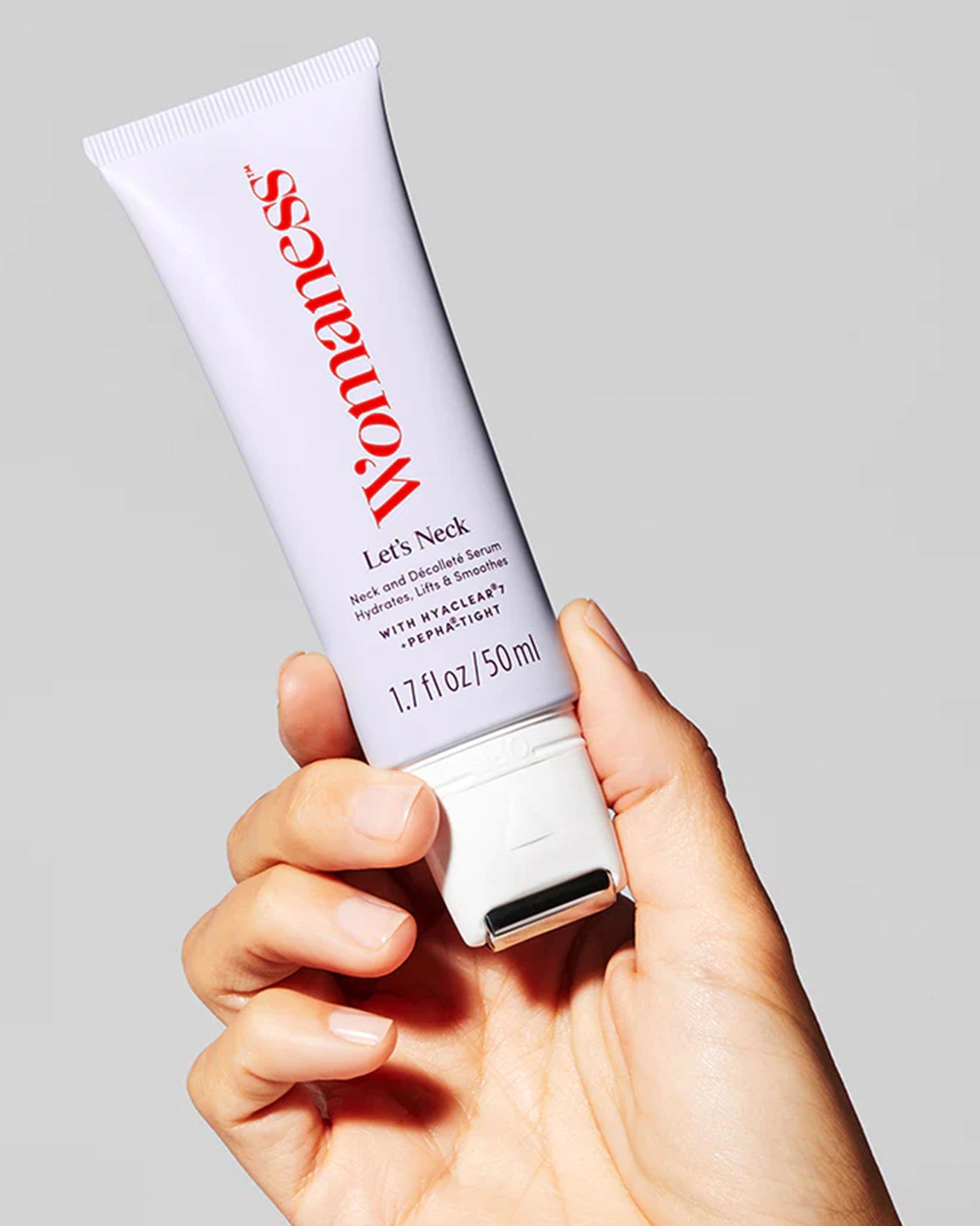 A hand holds a white tube labeled “Faire Let’s Neck – Neck Serum w/ Cooling Applicator” against a plain gray background, featuring red and black text and a cooling roller tip for neck and décolleté care.
