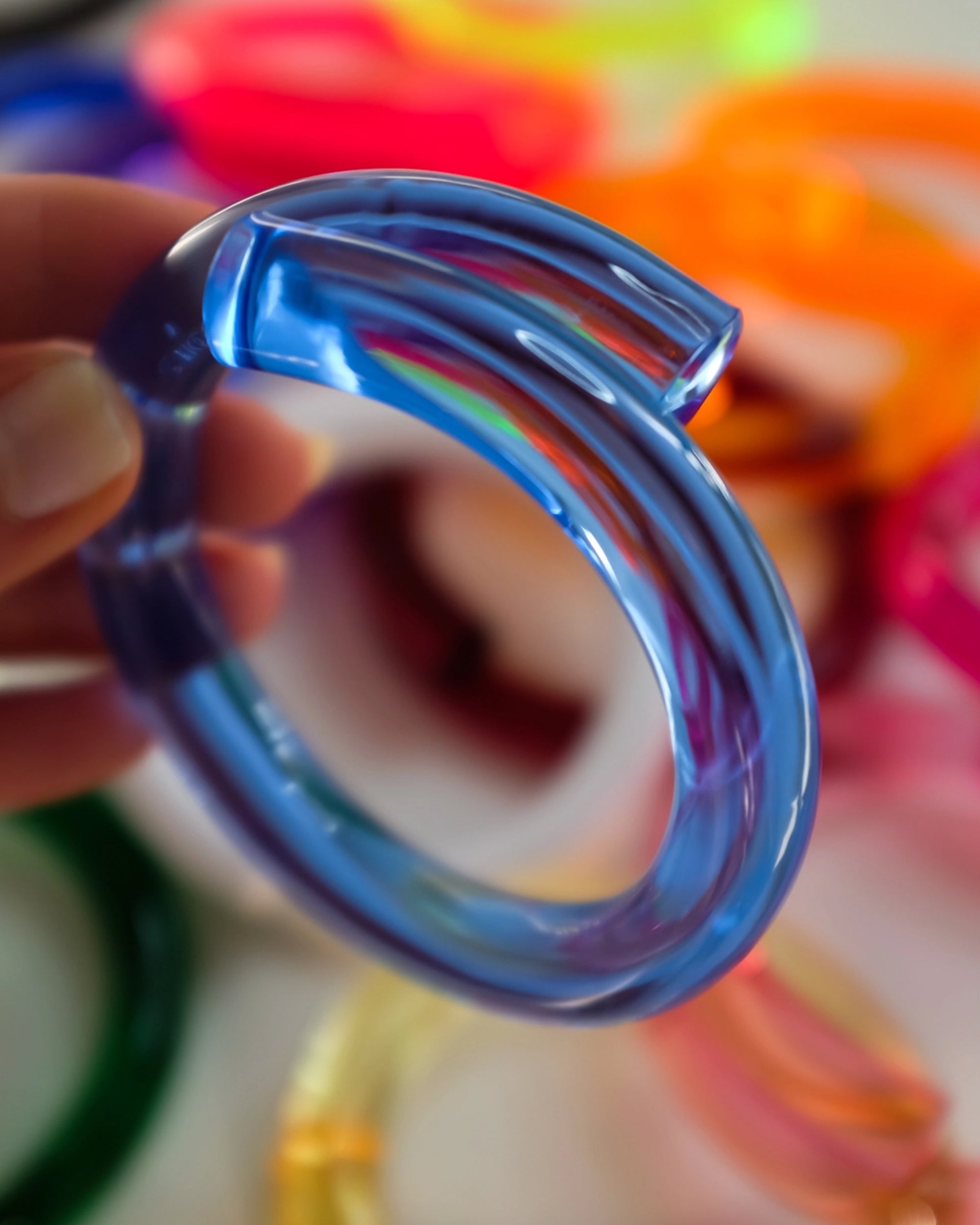 A hand holds the Faire Lucite Blue Neon Bracelet in the foreground, while several other colorful, minimalist jewelry pieces appear blurred in the background.