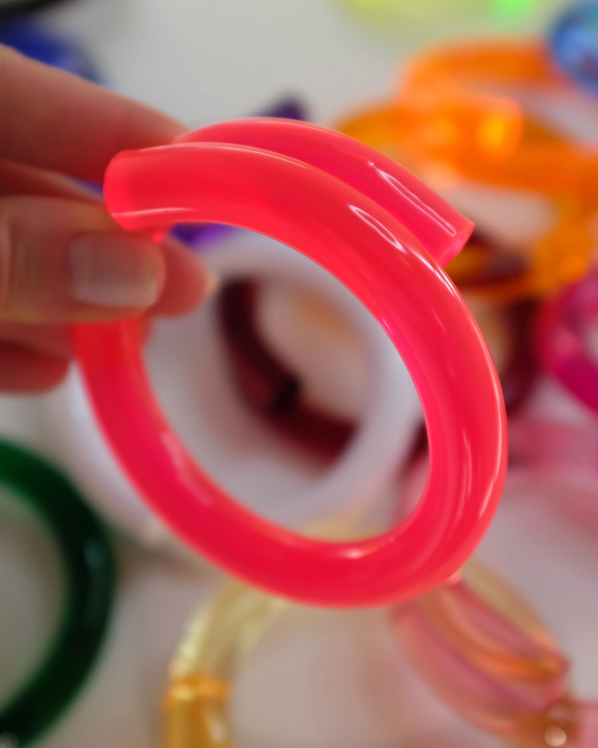 A hand displays the Faire Lucite Neon Bracelet in pink. Other vibrant Faire bracelets in orange, green, and yellow are softly blurred in the background.