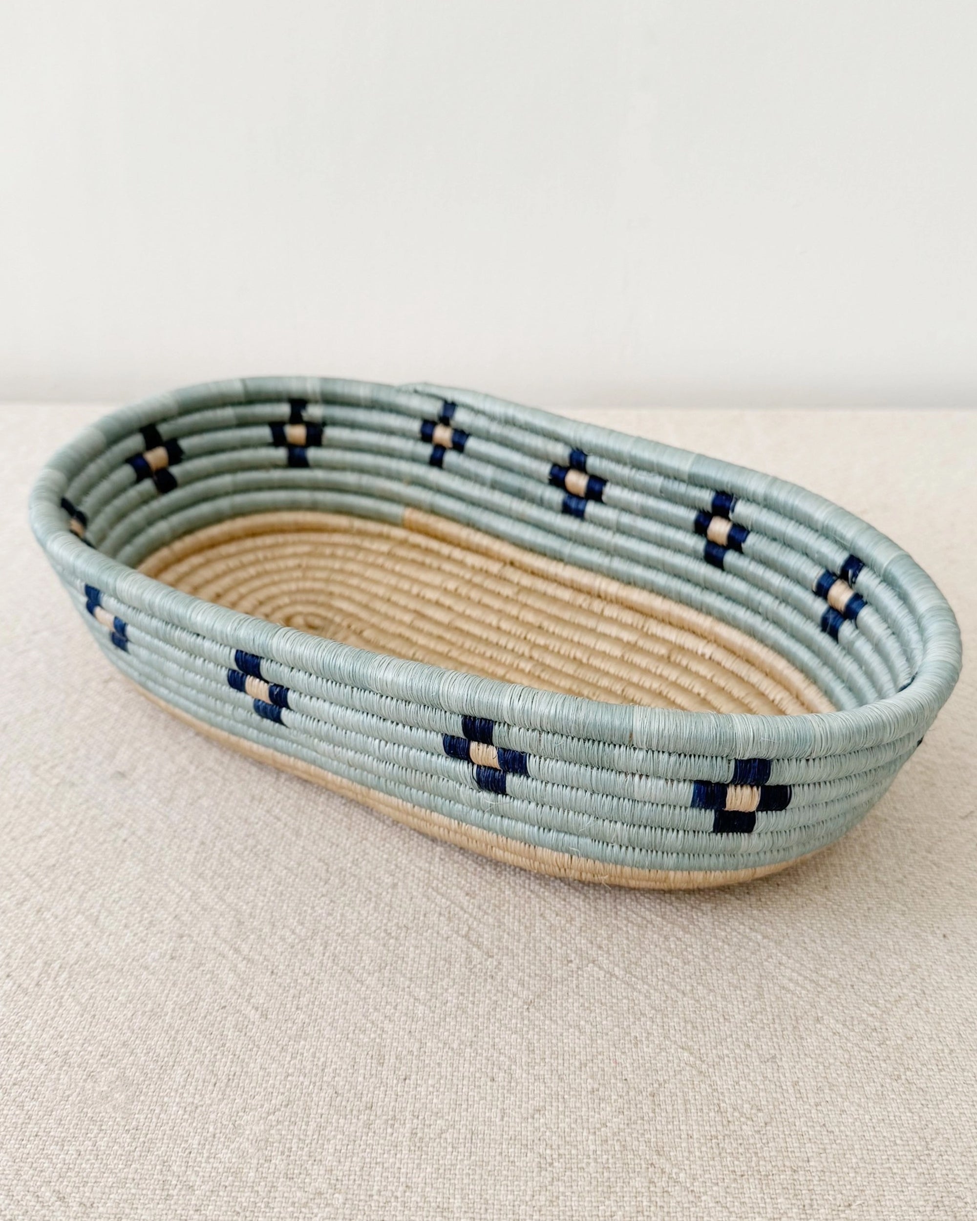 The Ngozi Bread Basket by Amsha - Faire is a shallow, oval woven basket with a light blue rim and sides, featuring small dark blue and cream square patterns. It rests on a textured beige surface.