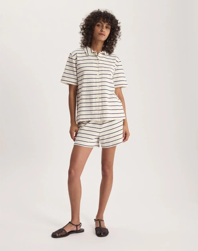 A person with curly hair wears the Nicia Shirt by Ines De LA Fressange—a cream short-sleeve striped button-front shirt—paired with matching black-striped shorts and black sandals, standing against a plain white background.