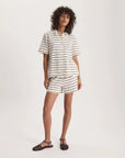 A person with curly hair wears the Nicia Shirt by Ines De LA Fressange—a cream short-sleeve striped button-front shirt—paired with matching black-striped shorts and black sandals, standing against a plain white background.