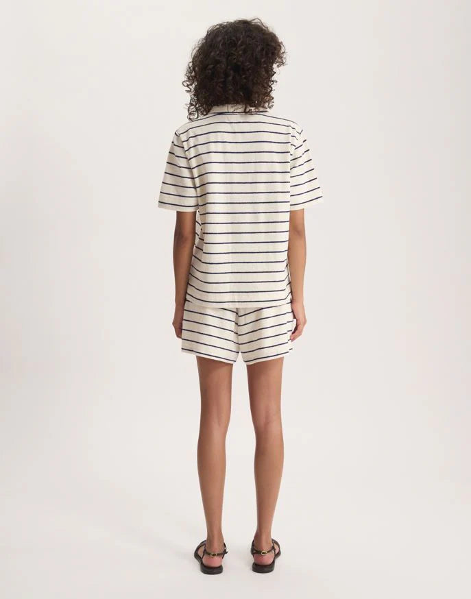 A person with curly hair faces away, wearing the Nicia Shirt by Ines De La Fressange— a short-sleeve, white, striped button-front shirt and matching shorts with black stripes—plus black sandals, against a plain white background.