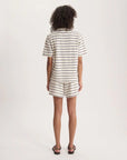A person with curly hair faces away, wearing the Nicia Shirt by Ines De La Fressange— a short-sleeve, white, striped button-front shirt and matching shorts with black stripes—plus black sandals, against a plain white background.