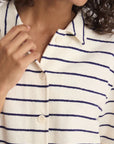 A person wearing the Nicia Shirt by Ines De LA Fressange—an organic cotton, striped button-front shirt with navy blue horizontal stripes and white buttons—touches the collar with one hand; their face is partially out of frame.