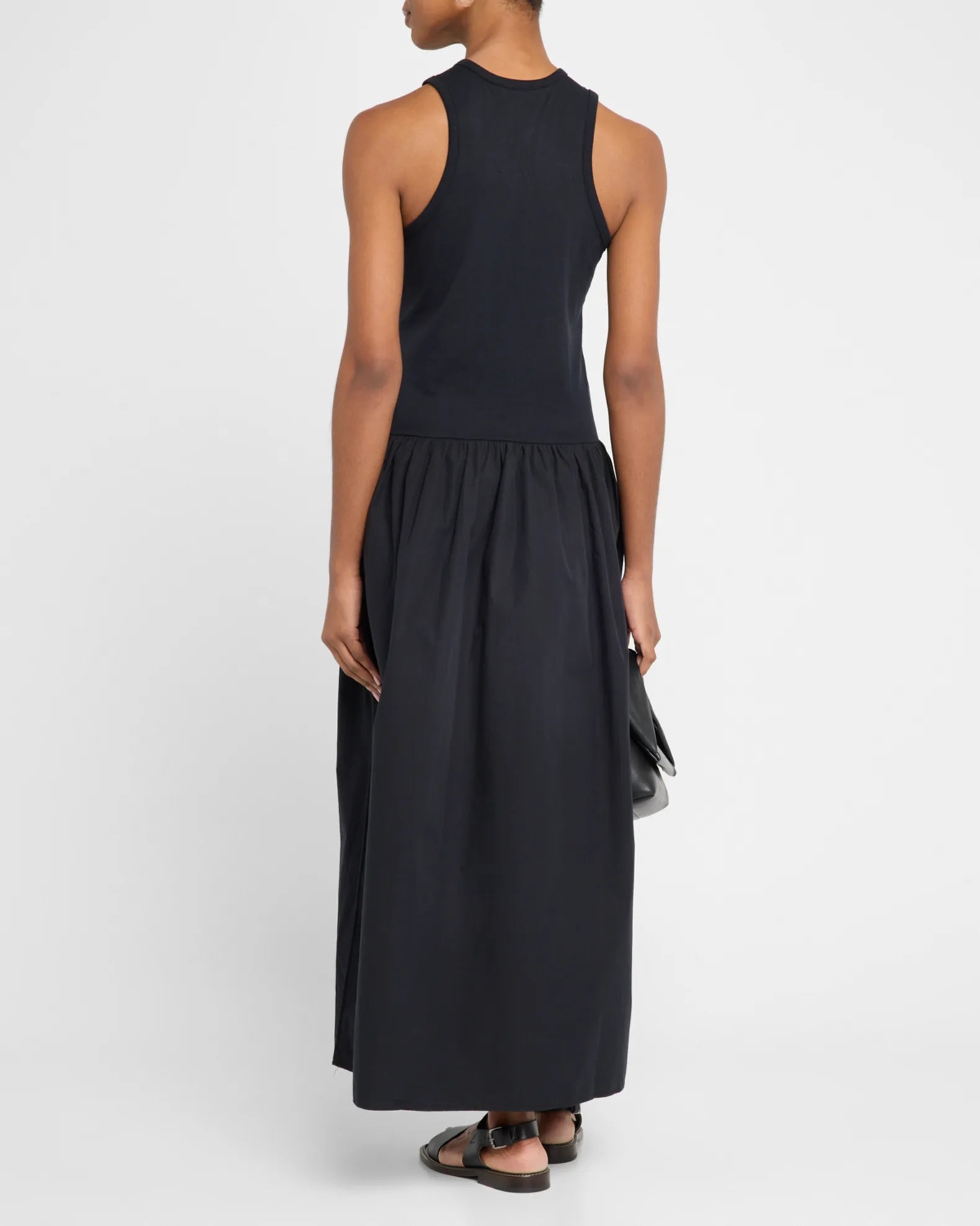 A person models the Racer Tank Dress by FRAME, standing with their back to the camera in a sleek, all-black ensemble. They hold a small clutch and wear sandals against a plain white background, perfectly capturing effortless style and simplicity.